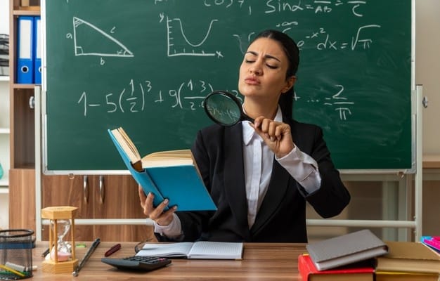 suspicious young female teacher sits table with school supplies reading book with magnifier classroom